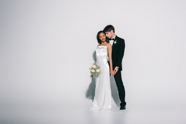 teljes hosszúságú kilátás elegáns vőlegény és gyönyörű afro-amerikai menyasszony fogja a kezét fehér háttér - Fotó, kép