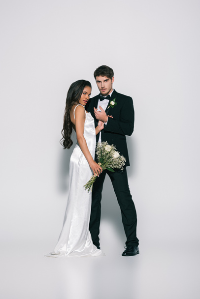 full length view of elegant bridegroom and attractive african american bride looking at camera while posing on white background - Fotoğraf, Görsel