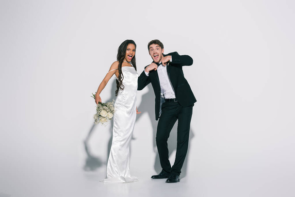 full length view of cheerful bridegroom touching bowtie near elegant african american bride on white background - Photo, Image