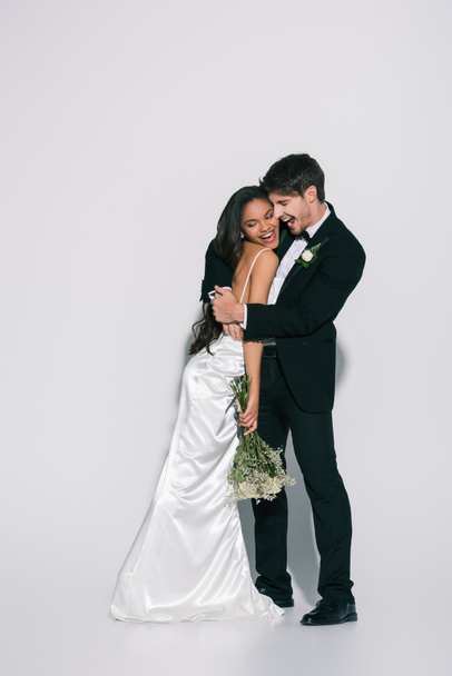 full length view of happy bridegroom embracing smiling african american bride on white background - Foto, Bild