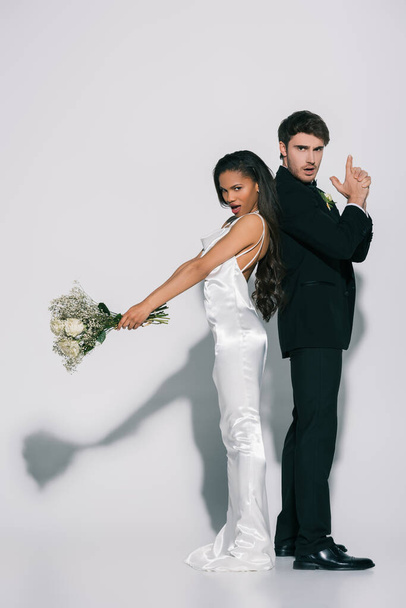 full length view of confident bridegroom showing gun gesture while standing back to back with african american bride with wedding bouquet on white background - Фото, зображення