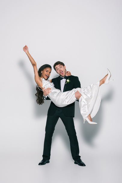 full length view of cheerful bridegroom holding happy african american bride on hands on white background - Zdjęcie, obraz