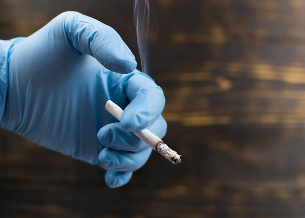 Cigarette smoking in a hand in a rubber glove on a dark background. - Foto, afbeelding