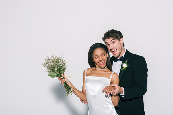 happy bridegroom showing wedding on hand of beautiful african american bride on white background - Photo, Image
