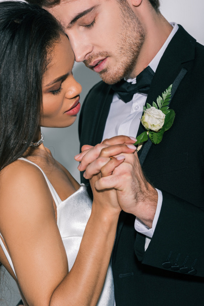 young interracial newlyweds holding hands while standing face to face with closed eyes on white background - Фото, изображение