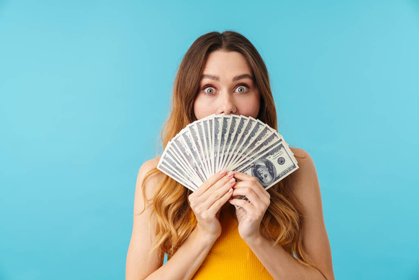 Portrait of beautiful joyous caucasian woman holding dollar banknotes isolated over blue background in studio - Фото, изображение