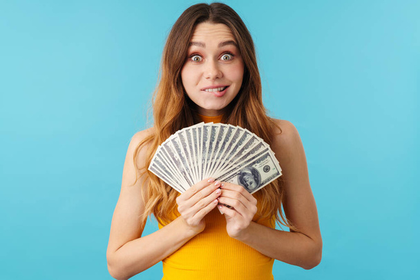 Portrait of beautiful joyous caucasian woman smiling and holding dollar banknotes isolated over blue background in studio - Foto, afbeelding