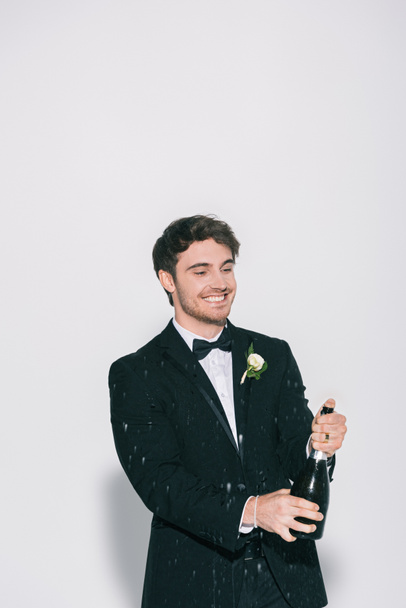 cheerful bridegroom opening bottle near splashes of champagne on white background - Photo, Image