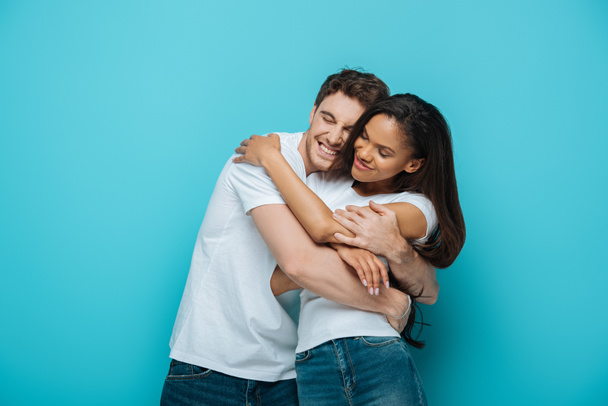 young, happy interracial couple in white t-shirts embracing with closed eyes on blue background - Photo, Image
