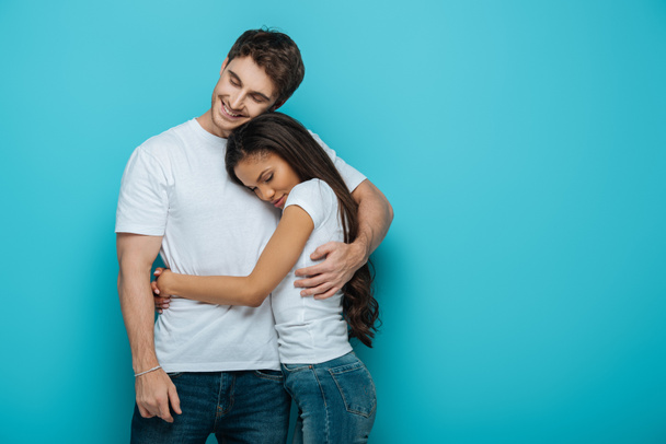 happy interracial couple embracing with closed eyes on blue background - Photo, Image