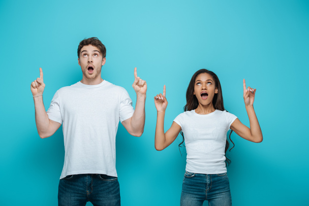 shocked interracial couple looking up and pointing with fingers on blue background - Foto, immagini
