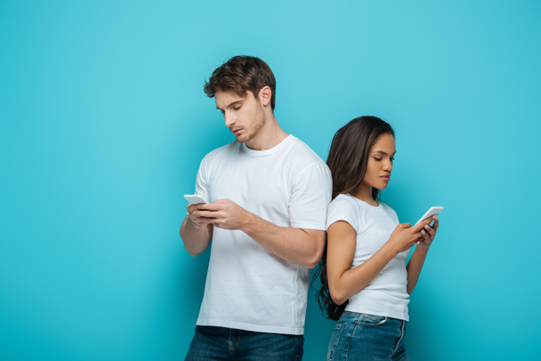 young interracial couple chatting on smartphones while standing back to back on blue background - Fotografie, Obrázek