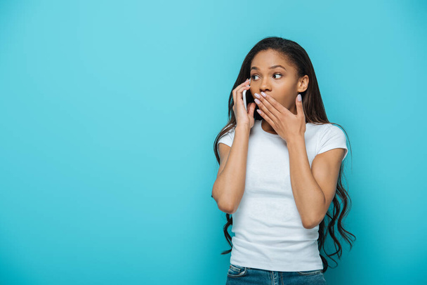 shocked african american girl covering mouth with hand while talking on smartphone isolated on blue - 写真・画像
