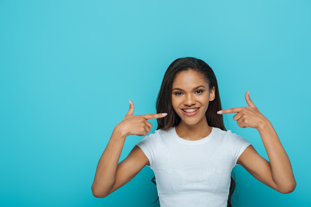 happy african american girl smiling at camera while pointing at braces on her teeth on blue background - Фото, изображение