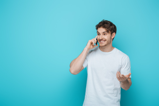 cheerful guy standing with open arm while talking on smartphone on blue background - Foto, Imagen