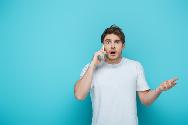 shocked man standing with open arm while talking on smartphone on blue background - Foto, Imagen