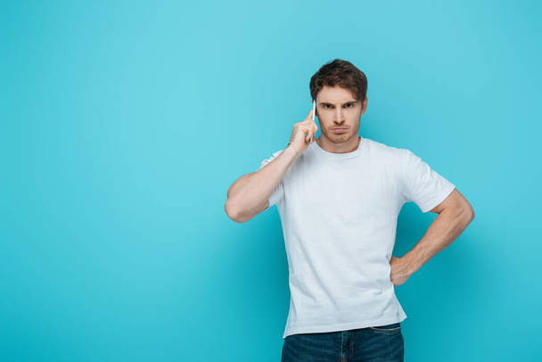 displeased man holding had on hip while talking on smartphone on blue background - Photo, Image