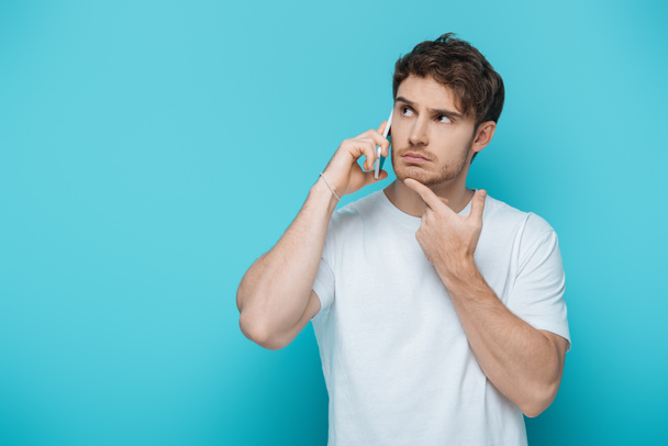 thoughtful guy touching chin while talking on smartphone and looking away on blue background - Foto, Imagem