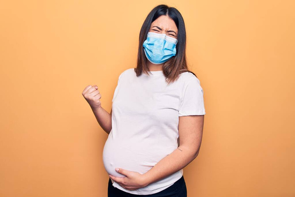 Giovane donna incinta madre di indossare maschera di protezione per la malattia coronavirica celebrando sorpreso e stupito per il successo con le braccia alzate e gli occhi chiusi
 - Foto, immagini