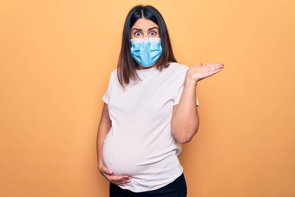Jeune femme enceinte mère de porter un masque de protection pour la maladie du coronavirus sans idée et confondue avec les bras ouverts, aucune idée et le visage douteux
. - Photo, image