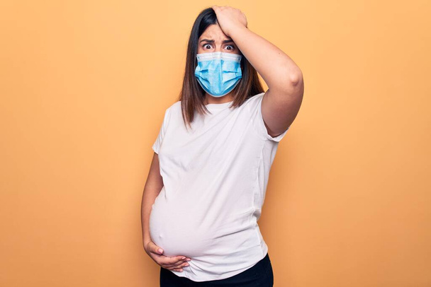 Jonge zwangere vrouw moeder te dragen beschermmasker voor coronavirus ziekte verrast met de hand op het hoofd voor fout, herinner fout. Vergeten, slecht geheugen concept. - Foto, afbeelding