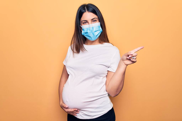 Mladá těhotná žena matka nosit ochrannou masku pro koronavirovou nemoc s veselým úsměvem ukazuje rukou a prstem až na bok - Fotografie, Obrázek