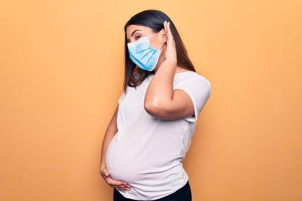 Jonge zwangere vrouw moeder draagt beschermmasker voor coronavirus ziekte glimlachen met de hand over oor luisteren en horen van geruchten of roddels. Doofheid. - Foto, afbeelding