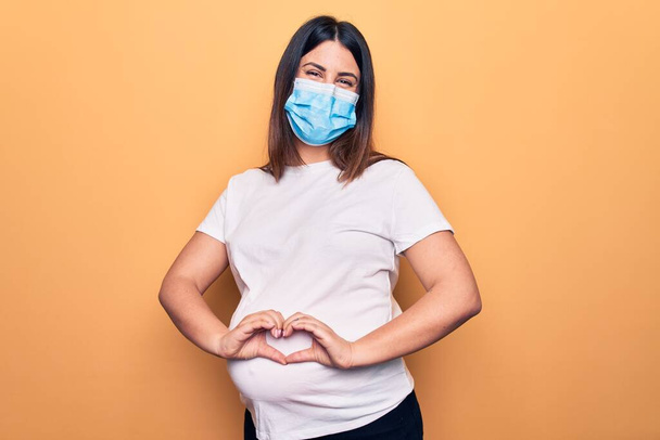 Jonge zwangere vrouw moeder te dragen bescherming masker voor coronavirus ziekte glimlachend in de liefde doen hart symbool vorm met de handen. Romantisch concept. - Foto, afbeelding