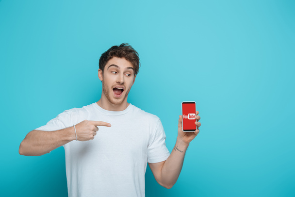 KYIV, UKRAINE - APRIL 23, 2019: excited guy pointing at smartphone with Youtube app on screen on blue background - Foto, Imagem