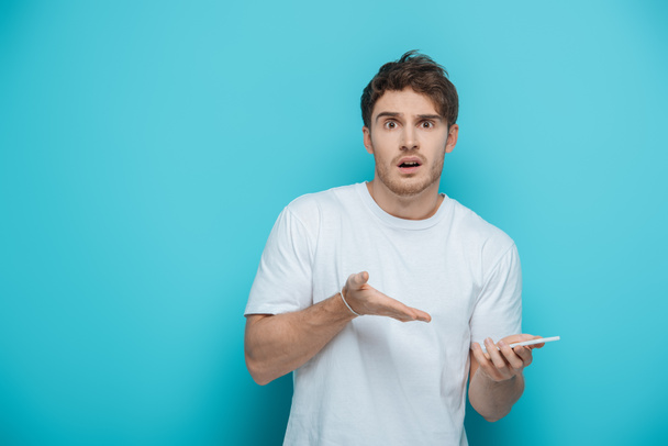 shocked young man pointing with hand at smartphone while looking at camera on blue background - Fotoğraf, Görsel