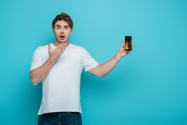 shocked guy holding hand near face while showing smartphone with business analytics app on blue background - Foto, Bild