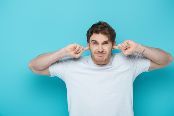 displeased young man plugging ears with fingers while looking at camera on blue background - Photo, Image
