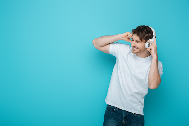 excited young man touching wireless headphones while listening music with closed eyes on blue background - Fotó, kép