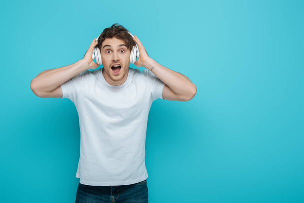 shocked young man touching wireless headphones while looking at camera on blue background - Fotografie, Obrázek