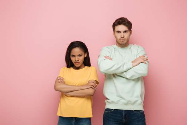 displeased interracial couple standing with crossed arms while looking at camera on pink background - Foto, imagen