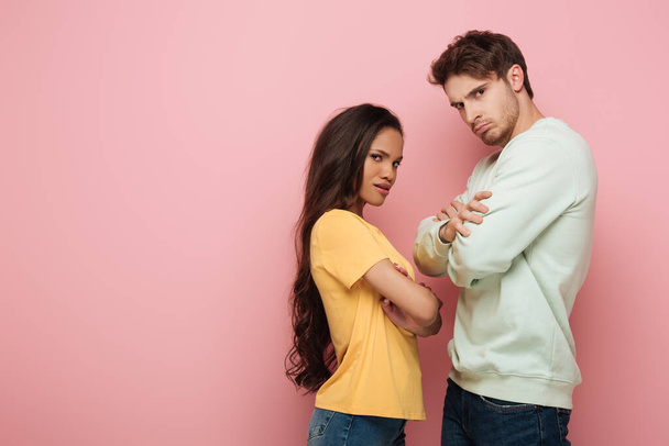 offended interracial couple standing with crossed arms while looking at camera on pink background - 写真・画像