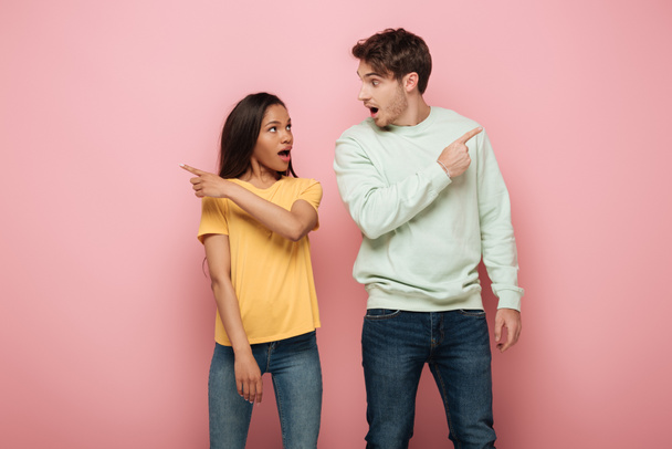 shocked interracial couple looking at each other and pointing away with fingers on pink background - Фото, зображення