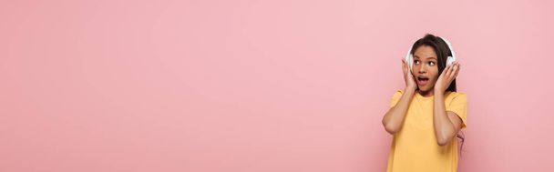 horizontal image of shocked african american girl touching wireless headphones while looking away isolated on pink - Photo, image