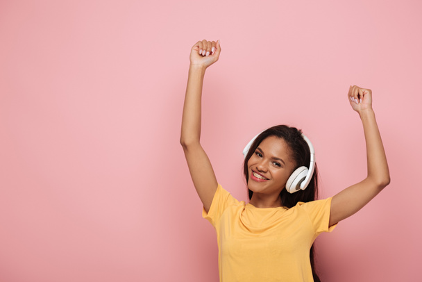 heureux afro-américaine fille dans casque sans fil danser avec les mains levées tout en regardant la caméra sur fond rose
 - Photo, image