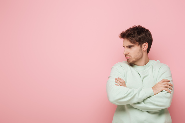 offended young man looking away while standing with crossed arms on pink background - Foto, Imagem