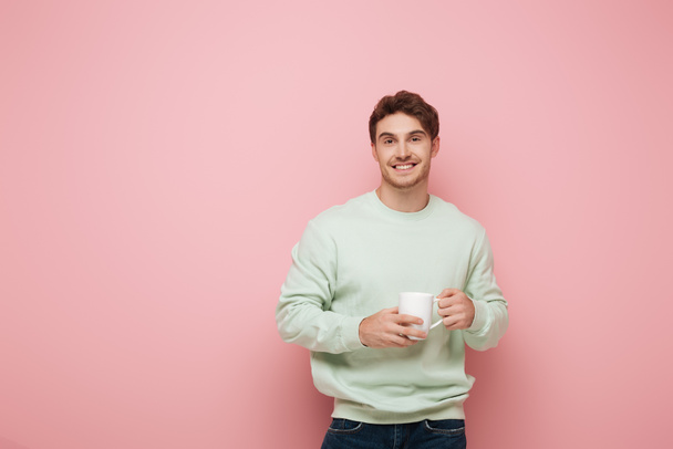 handsome young man holding white cup while smiling at camera on pink background - Foto, Imagem