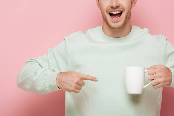 cropped view of smiling man pointing with finger at white cup on pink background - Foto, afbeelding