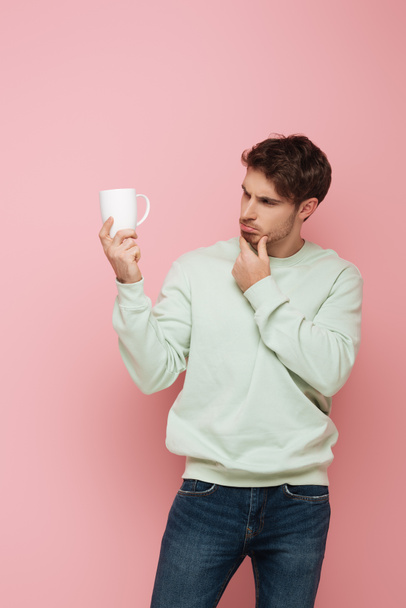 thoughtful guy touching chin while looking at white cup on pink background - Fotó, kép