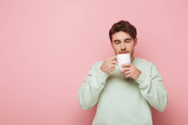 handsome young man enjoying flavor of coffee while holding white cup on pink background - Foto, immagini