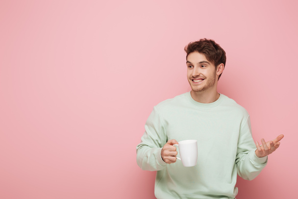 cheerful young man holding cup while standing with open arm and looking away on pink background - Fotografie, Obrázek