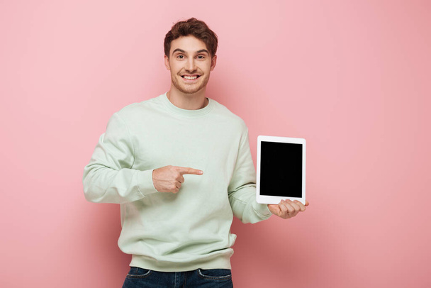 smiling man pointing with finger at digital tablet with blank screen while looking at camera on pink background - Foto, Bild