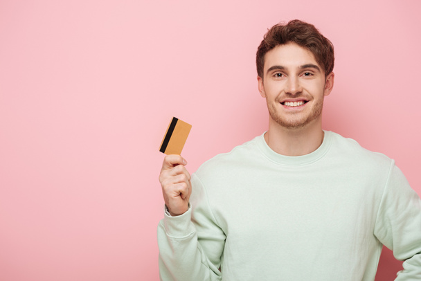 happy young man holding credit card while smiling at camera on pink background - Photo, Image