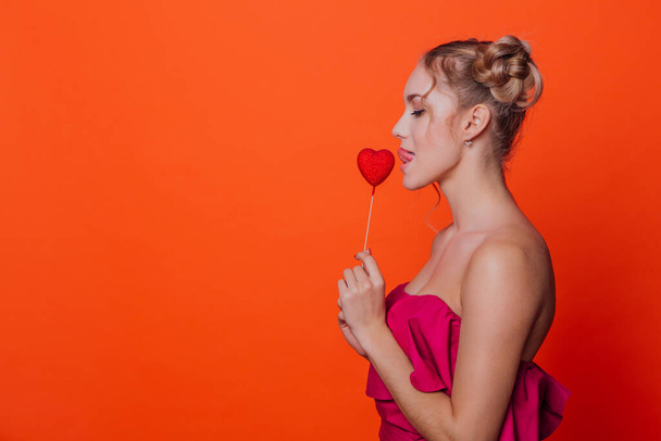 La chica en un vestido rojo sobre un fondo naranja en el estudio. Chica rubia con dos nudos de pelo sosteniendo el corazón rojo en el palo cerca de los labios. Copiar espacio
. - Foto, Imagen