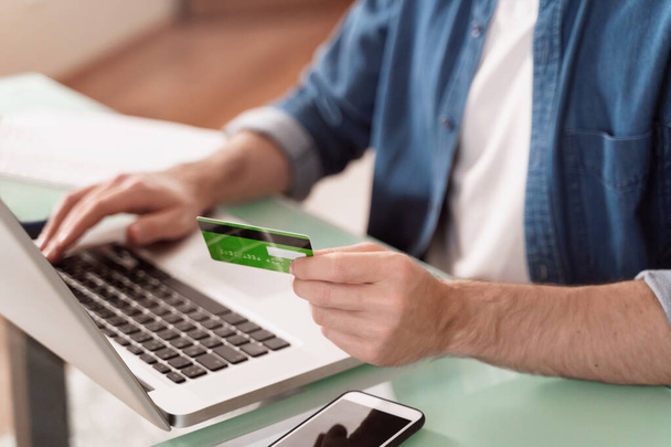 若い白人男性はオンラインで支払うためにクレジットカードとラップトップを使用しています。電子バンキング、電子商取引のためのガジェットを使用しています。商品のオンライン支払い。通貨交換のオンライン取引。電話でオンライン決済 - 写真・画像