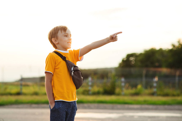 Kleine jongen die naar voren wijst. Leuk kind in geel t-shirt op een wandeling. Kind draagt trendy taille tas in de zomer dag. Jongen kijkt uit naar iets interessants. Knap gelukkig kind wijzend omhoog. - Foto, afbeelding